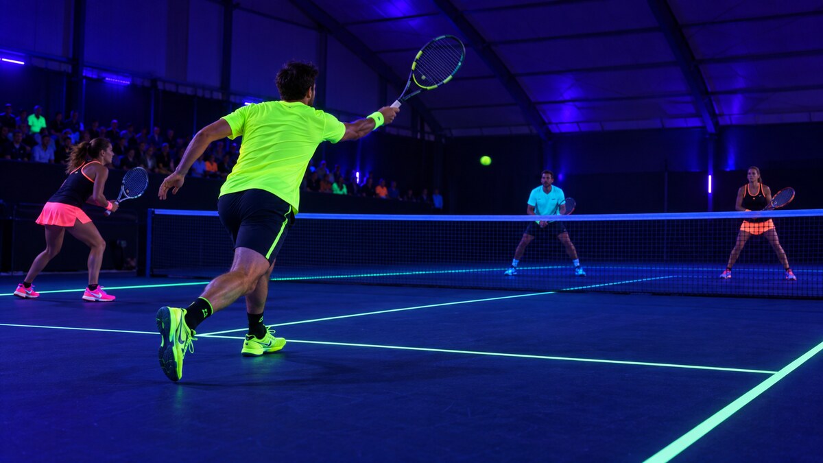Tournoi de tennis fluo en double sur un court couvert éclairé par lumière noire UV